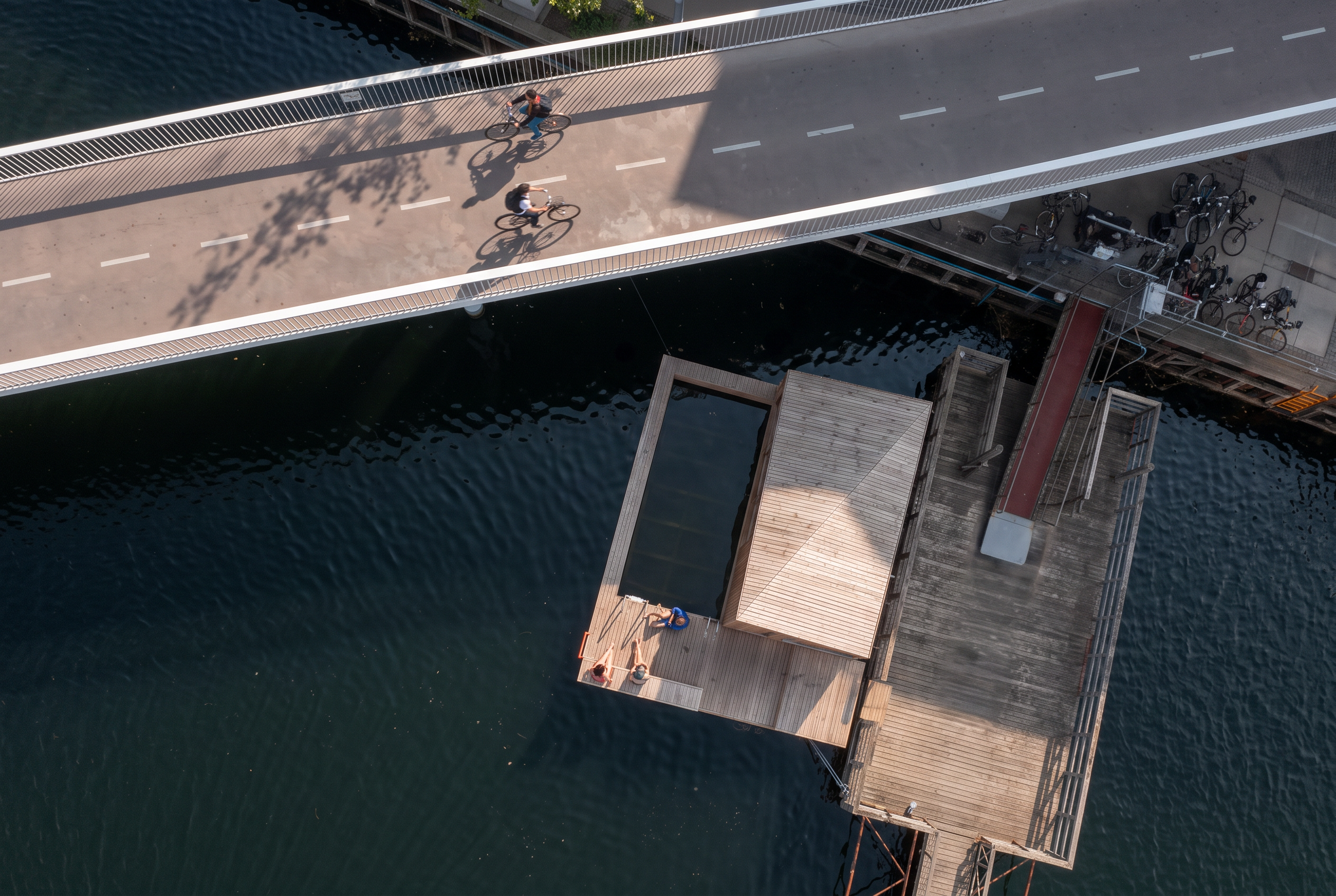 Stejlt luftfoto ovenfra af lys træsauna med åbent bassin på flydedæk, gæster på terrassen og cyklister på broen over havneløbet.